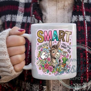 Könnte beinhalten: Weiße Keramik-Tasse mit farbenfroher Grafik. Das Design zeigt das Wort "SMART" in Gold und Leopardenmuster, einen Cartoon-Esel und den Text "FULL-TIME SMART-ASS". Zusätzlicher Text enthält "ZERO FILTER" und "WARNING OPINIONS MAY LEAK".