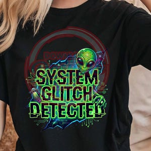Könnte beinhalten: Schwarzes T-Shirt mit einem grünen Alien-Design und dem Text "SYSTEM GLITCH DETECTOR". Die Grafik enthält ein UFO und Binärcode-Elemente, was ein Science-Fiction-Thema erzeugt.