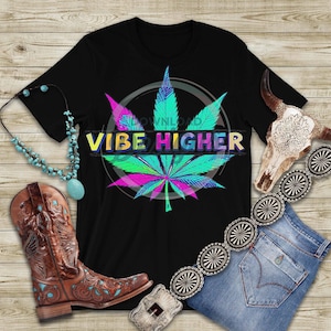 May include: Black t-shirt featuring a vibrant cannabis leaf design with the words "Vibe Higher." The image includes a turquoise necklace, brown cowboy boots, a decorative belt, a denim skirt, and a cow skull.