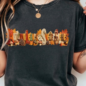 Könnte beinhalten: Ein anthrazitfarbenes Kurzarm-T-Shirt mit einem Grafikdruck und dem Text Autumn Leaves & Sweater Sleeves. Das Design ist mit buntem Herbstlaub, Tannenzapfen, Eicheln und kleinen Pilzen in verschiedenen Orange- und Brauntönen verziert.