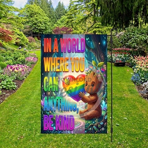 Könnte beinhalten: Eine Gartenflagge mit dem Text "IN A WORLD WHERE YOU CAN ANYTHING, BE KIND" in Regenbogenfarben. Die Flagge zeigt eine Zeichentrickfigur, die ein Regenbogenherz hält, vor einem Waldhintergrund.