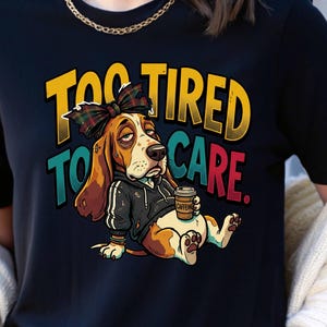 Könnte beinhalten: Schwarzes T-Shirt mit einem Cartoon-Basset-Hound mit karierter Schleife und Kapuzenpullover, der eine Kaffeetasse hält. Der Text lautet "TOO TIRED TO CARE" in Gelb, Türkis und Rot. Der Hund hat einen müden Gesichtsausdruck.