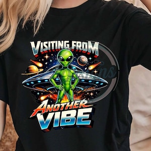 Könnte beinhalten: Schwarzes T-Shirt mit einem grünen Alien vor einem UFO. Der Text "VISITING FROM ANOTHER VIBE" ist in einer farbenfrohen, futuristischen Schriftart dargestellt. Das Design umfasst Planeten und Sterne.