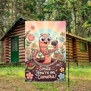 Könnte beinhalten: Eine Gartenflagge mit einem Cartoon-Wurm, der eine Kamera hält, mit dem Text "Smile... You're on Camera!". Die Flagge hat ein farbenfrohes Design mit Blumen und einem Marienkäfer, vor dem Hintergrund einer Blockhütte und Bäumen.