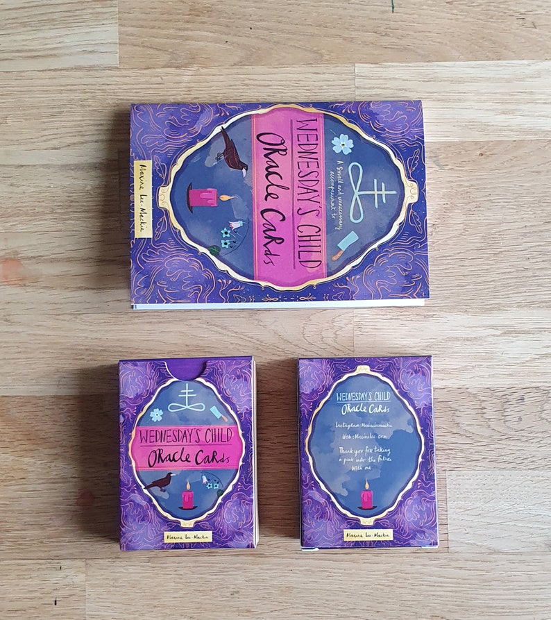 May include: A set of oracle cards with a purple and gold design. The cards feature a black crow, a candle, and a symbol. The box for the cards has the text "Wednesday's Child Oracle Cards" on the front.