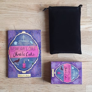 May include: A purple and gold box with the text "Wednesday's Child Oracle Cards" and a small illustration of a bird, candle, and flowers. The box is sitting on a wooden surface next to a black velvet bag.