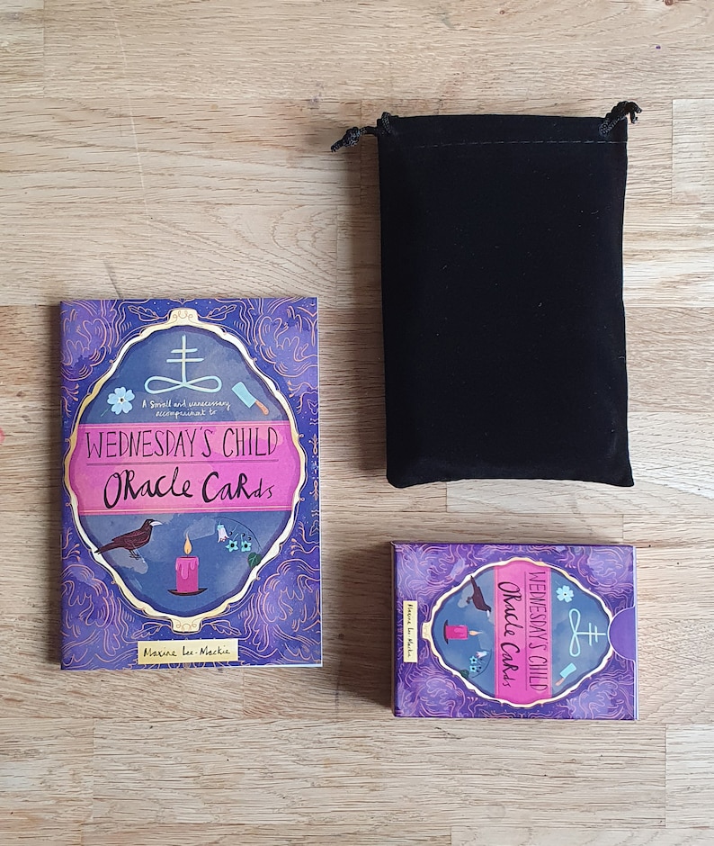 May include: A purple and gold box with the text "Wednesday's Child Oracle Cards" and a small illustration of a bird, candle, and flowers. The box is sitting on a wooden surface next to a black velvet bag.