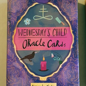 May include: A purple box with a gold border and a pink and blue illustration of a raven, a candle, and a knife. The text on the box reads "Wednesday's Child Oracle Cards" and "Maxine Lee Mackie".