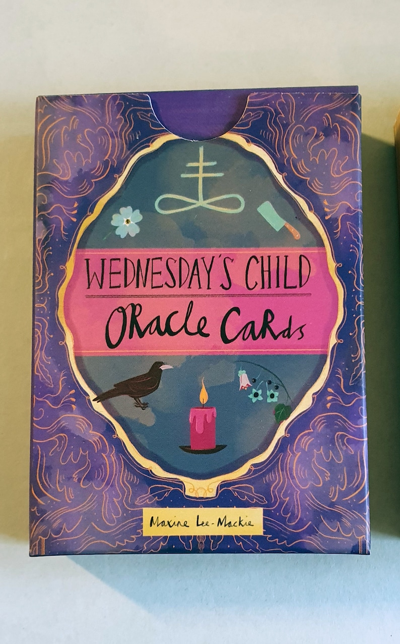 May include: A purple box with a gold border and a pink and blue illustration of a raven, a candle, and a knife. The text on the box reads "Wednesday's Child Oracle Cards" and "Maxine Lee Mackie".