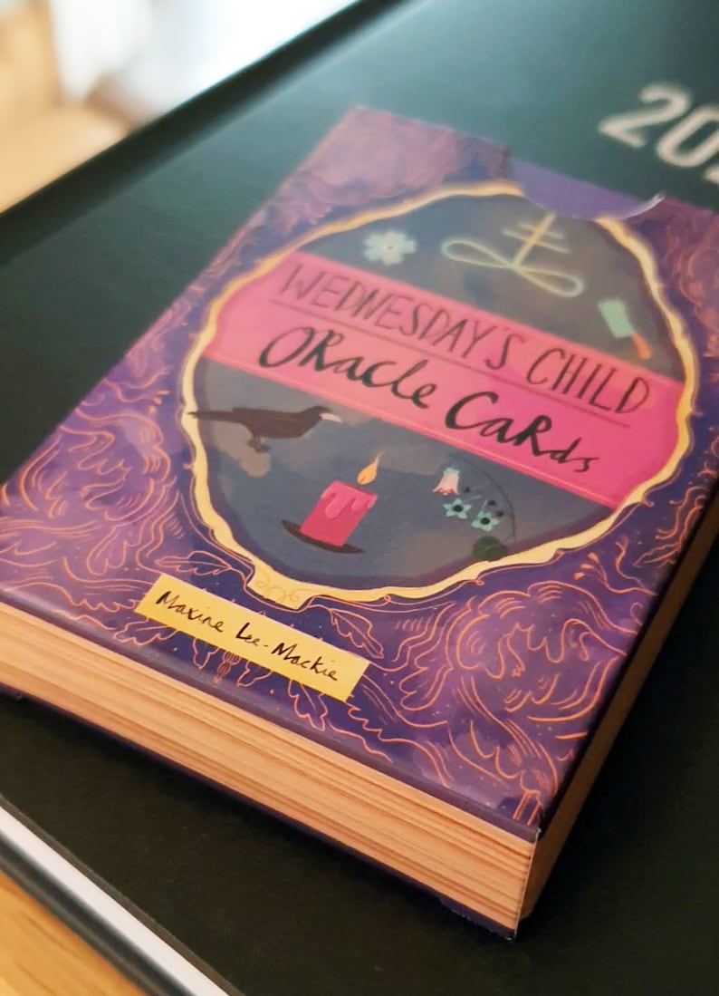 May include: A purple and gold deck of oracle cards with the title "Wednesday's Child Oracle Cards" and the author's name "Maxine Lee Mackie".