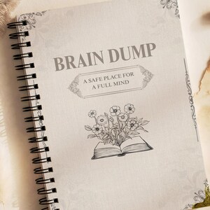 Peut inclure: Un carnet à reliure spirale avec une couverture beige clair portant le texte BRAIN DUMP et A SAFE PLACE FOR A FULL MIND. La couverture présente une illustration au trait noir de fleurs sortant d'un livre ouvert et des ornements décoratifs.