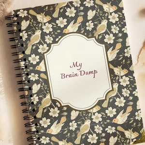 May include: A spiral-bound notebook with a dark grey cover featuring a repeating pattern of light-coloured birds and flowers. A central cream-coloured label with a decorative border displays the dark red text My Brain Dump in the centre.