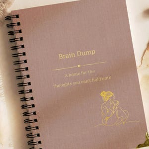 Może przedstawiać: Brudnoróżowy notatnik ze spiralą, ze złotym napisem Brain Dump, A home for the thoughts you can't hold onto, oraz złotą grafiką liniową przedstawiającą siedzącą osobę trzymającą kubek, wykończony ozdobnym metalowym narożnikiem.