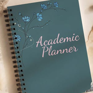 May include: A teal spiral-bound academic planner featuring a delicate illustration of blue flowers and thin black stems on the left side. The text "Academic Planner" is printed in a light pink script font in the centre. The cover has silver corner accents.