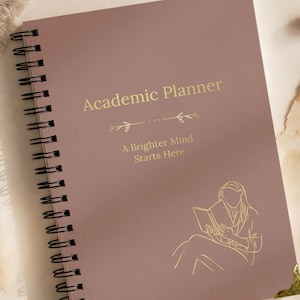 May include: A dusty rose spiral-bound academic planner featuring gold foil text that reads Academic Planner and A Brighter Mind Starts Here, with a gold line art illustration of a person reading a book and a decorative gold metal corner protector.
