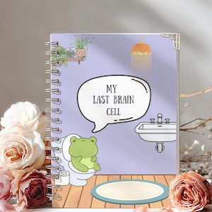 May include: A spiral-bound notebook with a light purple cover featuring a cartoon green frog sitting on a toilet. The cover includes a speech bubble with the text MY LAST BRAIN CELL, a sink, a hanging plant, and a light fixture above a wooden floor.
