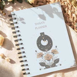 Może przedstawiać: Jasnoniebieski planer w czarnej oprawie spiralnej. Na okładce znajduje się napis TIANA'S PLANNER, ciemna okrągła grafika niebiańska z półksiężycem oraz szczegółowa ilustracja kwiatów z ciemnozielonymi liśćmi.
