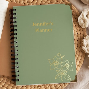 May include: A sage green spiral-bound planner with black wire binding. The cover features the gold text "Jennifer's Planner" and a gold line-art floral illustration. Decorative gold metal corner protectors are attached to the top and bottom right.