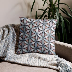 May include: A decorative throw pillow with a geometric floral pattern. The design features overlapping petal shapes in shades of blue, pink, and gray, creating a symmetrical and visually appealing design. The pillow is square and rests on a patterned blanket.