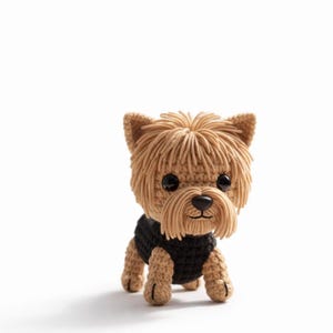 Könnte beinhalten: Ein gehäkelter Yorkshire Terrier Hund mit hellbraunem Fell und einem schwarzen Pullover. Der Hund hat schwarze Augen und eine schwarze Nase. Der Hund steht auf einer weißen Oberfläche.