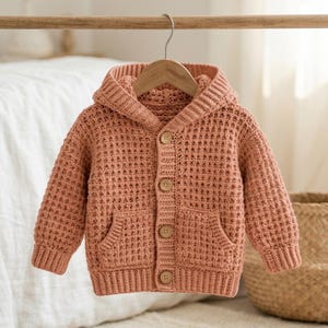 Könnte beinhalten: Eine altrosa langärmelige Strickjacke mit Kapuze, Waffelstrickmuster, einer Knopfleiste mit vier hellbraunen Holzknöpfen und zwei aufgesetzten Vordertaschen. Das Kleidungsstück verfügt über gerippte Ärmelbündchen und einen gerippten Saum.