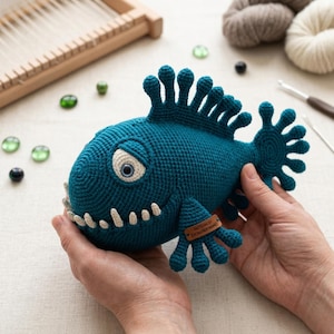 Könnte beinhalten: Ein gehäkeltes Fischspielzeug in Türkis mit weißen Zähnen, einem beigen Auge und einem Etikett mit der Aufschrift "PATTERN BY DESIGNER NAME". Der Fisch hat ein einzigartiges Design mit flossenartigen Anhängen.