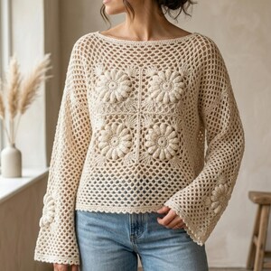 Könnte beinhalten: Ein cremefarbener, langärmeliger Häkelpullover mit weitem U-Boot-Ausschnitt und einem durchgehenden Netzmuster. Das Vorderteil ist mit vier großen, erhabenen Blumenmotiven verziert, während sich weitere florale Akzente an den Ärmeln befinden.