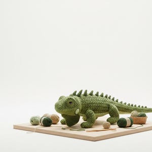 May include: A handmade, green crochet iguana with detailed features, including a textured body and spiky dorsal scales. The iguana is displayed on a wooden board with yarn balls and a small basket. The iguana measures approximately 18 inches in length.