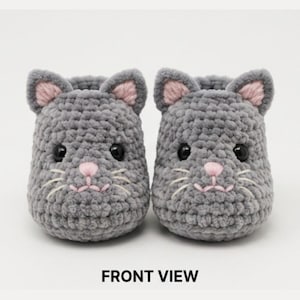 Könnte beinhalten: Ein Paar graue Häkelschuhe in Form von Katzenköpfen mit rosa Innenohren, rosa gestickten Nasen und weißen gestickten Schnurrhaaren. Die Schuhe haben schwarze Sicherheitsaugen aus Kunststoff und eine strukturierte Oberfläche. Text FRONT VIEW.
