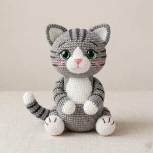 Könnte beinhalten: Ein gehäkeltes Stofftier in Form einer Katze aus grauem und weißem Garn. Die Katze hat dunkelgraue Streifen auf Kopf und Schwanz, große grüne Augen, eine rosa gestickte Nase sowie weiße Flecken an Brust, Pfoten und den Innenseiten der Ohren.