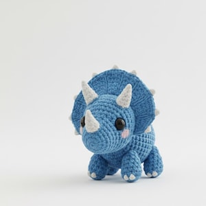 Könnte beinhalten: Ein blau gehäkeltes Triceratops-Dinosaurier mit weißen Hörnern und Stacheln. Das handgefertigte Plüschtier hat große schwarze Augen und rosa Wangen, was ihm ein freundliches Aussehen verleiht. Der Dinosaurier steht auf einer weißen Oberfläche.