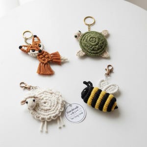 Könnte beinhalten: Vier handgefertigte Häkel-Schlüsselanhänger: ein Fuchs mit orangefarbenen und weißen Details, eine grüne Schildkröte, ein weißes Schaf und eine schwarz-gelbe Biene. Jeder Schlüsselanhänger hat einen Metallverschluss und einen Ring. Der Schaf-Schlüsselanhänger hat ein Etikett mit der Aufschrift "Handmade with Love The Cozy Hook."