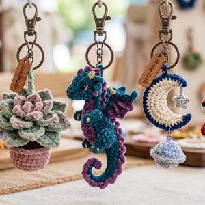 Könnte beinhalten: Fünf Häkel-Schlüsselanhänger an Metallverschlüssen. Die Designs umfassen einen dunkelblauen Halbmond mit Stern, eine grüne Sukkulente im rosa Topf, einen blaugrünen und violetten Drachen, einen cremefarbenen Mond mit Planet und einen orangefarbenen Fuchs mit einem roten Pilz.