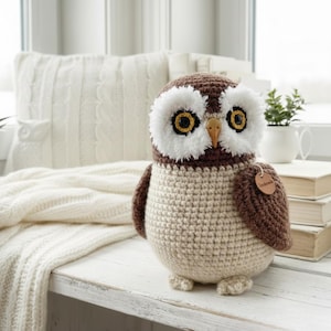 Könnte beinhalten: Eine handgefertigte Häkel-Eule in Braun-, Creme- und Weißtönen. Die Eule hat große, runde Augen mit gelben Zentren und einen kleinen Holzschnabel. Ein kleines Etikett mit der Aufschrift "Handmade" ist am Flügel der Eule befestigt. Die Eule sitzt auf einer weißen Oberfläche.