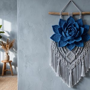 Könnte beinhalten: Ein Wandbehang mit einer großen, tiefblauen gehäkelten Lotusblume über einem komplexen Makramee-Design aus hellgrauer Kordel. Das gesamte Stück ist an einem horizontalen Holzast befestigt, der oben eine Schlaufe aus geflochtener Kordel besitzt.