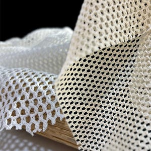 May include: Two pieces of white mesh fabric with different sized holes. The fabric is folded and draped over a wooden surface.