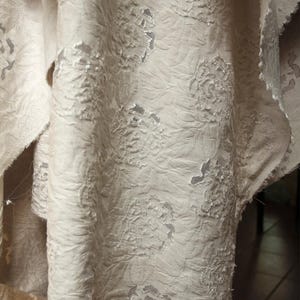 Puede incluir: Primer plano de una tela blanca con un patrón floral en relieve. La tela está doblada y drapeada, mostrando la textura del diseño.