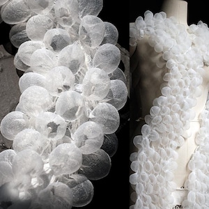 Puede incluir: Una prenda de vestir blanca translúcida hecha de tela con muchas formas pequeñas, redondas y con forma de burbuja. La prenda está drapeada sobre un maniquí.