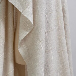 May include: Close-up of a cream-colored woven fabric with a textured, grid-like pattern. The fabric is draped, showing folds and the raw edge of the material. The neutral color and texture suggest it could be used for home decor or apparel.