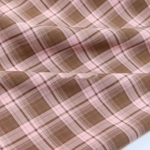 Könnte beinhalten: Nahaufnahme eines braunen und rosa karierten Stoffes. Der Stoff hat ein klassisches Karomuster mit sich kreuzenden Linien in Braun- und Rosatönen. Der Stoff ist gefaltet, wodurch Schatten und Textur entstehen.