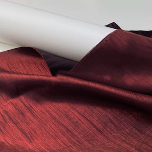 Op de afbeelding: Een close-up van een rol wit papier naast een dieprode stof. De stof heeft een getextureerde, licht iriserende uitstraling, met diagonale lijnen. De kleur is een rijke, donkere tint rood, met hints van bordeaux.