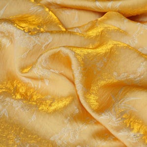 May include: Close-up of a luxurious, shimmering fabric in shades of gold and yellow. The fabric has a textured surface with subtle floral patterns in a lighter hue, creating a sense of depth and elegance. The folds and drapes of the material add to its rich appearance.