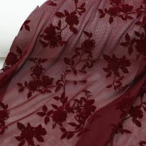 May include: Burgundy floral lace fabric. The fabric has a sheer mesh base with raised, velvety floral patterns. The flowers and leaves are in a darker shade of burgundy, creating a subtle contrast. The fabric is draped, showing its texture and flow.