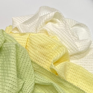 May include: Close-up of three fabrics in shades of yellow and white. The fabrics have a textured, grid-like pattern. The colors range from a pale, creamy white to a bright, sunny yellow, with a hint of green. The fabrics are draped and folded.