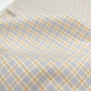 May include: Close-up of a fabric with a plaid pattern. The fabric has a light grey background with a grid of intersecting lines in light brown and yellow. The fabric is slightly wrinkled, creating soft shadows.