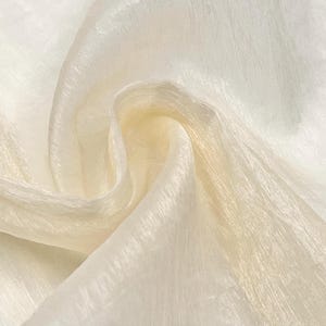 Op de afbeelding: Close-up van een vloeiende, gebroken witte stof met een subtiele glans. Het materiaal is delicaat gedrapeerd en toont zijn textuur en lichtreflecterende eigenschappen. De stof lijkt zacht en lichtgewicht, ideaal voor kleding of decoratieve projecten.