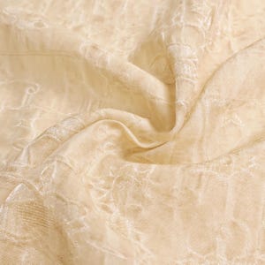 May include: Close-up of a beige fabric with a delicate, embroidered floral pattern. The fabric is draped, showing soft folds and subtle variations in color and texture. The overall effect is one of elegance and refinement.
