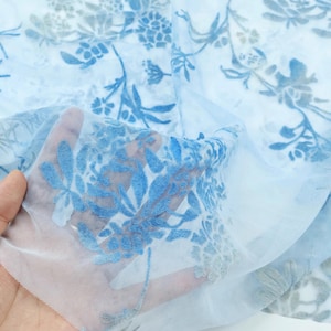 May include: Light blue floral patterned fabric. The sheer material features a delicate design of blue and gray flowers and leaves. The fabric is held up, showing its transparency and intricate details.