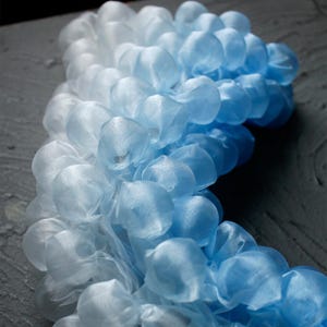 May include: A close-up of a decorative garland in shades of blue and white. The garland is made of small, gathered fabric puffs, creating a textured, cloud-like appearance. The gradient effect transitions from white to light blue to a deeper blue.