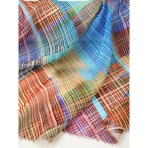 May include: A colorful scarf with a plaid pattern in shades of blue, orange, brown, and green. The fabric is lightweight and flowing, with a soft texture. The scarf is folded and draped, showcasing the vibrant color palette and design.